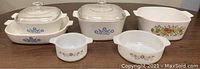 Front view of five Corning Ware casserole dishes with lids and different vintage patterns on a brown surface.
