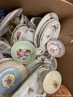 Top view of assorted plates, cups, and saucers with floral designs and gold trim, showing various pastel colors and patterns.