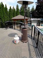 Full view of brown propane patio heater standing on deck next to white propane cylinder and pool in background