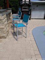 Three Hampton Bay outdoor high stools with silver metal frames and blue-green fabric seats beside stone outdoor counter and grill area.