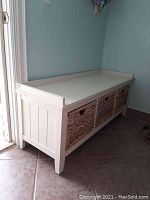 Wooden white bench with three woven wicker basket drawers underneath, placed against a wall on tiled floor.