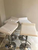Four white adjustable bar stools with chrome metal bases arranged in a corner showing the seats and base.