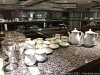 Wide view of espresso cups and saucers, creamer and sugar bowl, coffee pots, and silver rimmed goblets on marble countertop in mirrored background.