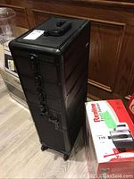 Black rolling makeup case standing upright, showing height and design with metal latches on the front and handle on top.