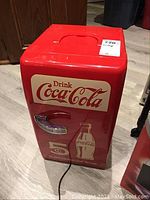 Front view of red Coca Cola mini fridge cooler with handle, logo, and bottle graphic. Power cord visible at bottom.