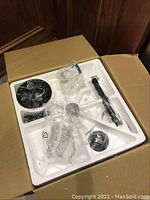 Unassembled fan parts in original box: mounting bracket, downrod, cage, motor housing, blade assembly components in styrofoam tray