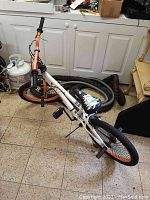 Full view of 20 inch Mongoose Crush bike showing white frame with orange rims and handlebars, decorative padded black and white seat, front and rear foot pegs, kickstand, and knobby tires.