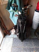 Photo of golf bag with several golf clubs standing on tiled floor near a door with blue cloth hanging behind.