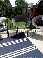 Two round back resin wicker chairs with black metal legs on outdoor upper deck, sitting on concrete with railing and outdoor carpet visible.