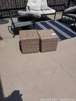 Two square wicker style foot stools placed on a patio floor next to outdoor seating furniture.
