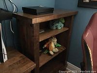 Photo showing the entire solid wood bookcase with two shelves and plush toys on the shelves, positioned next to a red chair.
