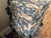 Stack of 15 blue and beige floral patterned seat cushions viewed from side angle showing thickness and ties.