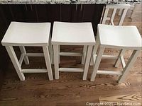 Three white wooden bar stools seen from the front, showing square seats, straight legs, and horizontal supports.