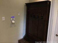 Front view of dark wood hall tree against light colored wall, showing four black metal hooks on upper backing panel and bench with storage below.