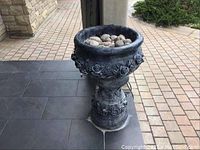 Full view of the large dark gray outdoor planter showing floral relief design, filled with stones, placed on tiled porch.
