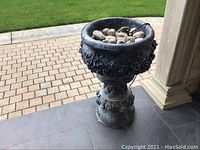 Full view of the dark gray heavy cement planter with decorative rose motifs filled with dirt and stones, showing the overall shape and setting outdoors.