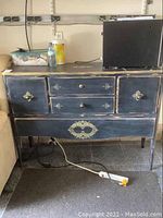 Front view of the wood cabinet showing decorative metal hardware, two side doors, two drawers with one knob missing, and an ornate metal lower panel decoration. Cabinet has distressed dark blue paint and visible scratches.