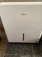 Front view of white Midea dehumidifier on dark floor showing Midea logo and water level indicator window