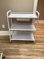 Full view of white metal frame cart showing three shelves and a drawer under the top shelf, on caster wheels.