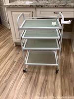 Front angled view of white metal framed cart with 4 clear glass shelves and clamp-on side attachment, on wooden floor.