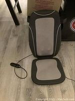 Full view of the black and gray massage and heated seat laid flat on a wood floor, showing wired remote control attached.