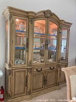 Front view of large lighted china cabinet with ornate crown molding, glass doors, and wooden base cabinetry. Contents inside are visible but not included.