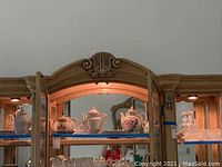 Porcelain teapots, sugar bowls and clear glass decanters inside lit wooden cabinet.
