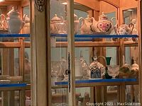 Frontal photo of china cabinet glass shelves showing porcelain teapots, clear glass dessert bowls, pitchers and decorative glassware arranged on two shelves.