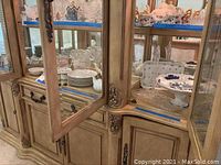 Lower shelf of display cabinet showing floral patterned plates, crystal ice bucket, teapots, and small serving dishes arranged neatly