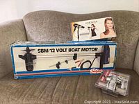 Overall view of the boxed Sevylor SBM 12 Volt boat motor, boxed Hair Made hair dryer stand, and packaged Kwikset door handle set arranged on couch.