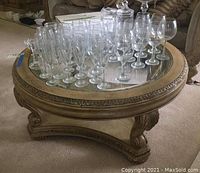 Angle view of round wood coffee table with detailed carved legs and circular glass top inlay. Various wine and liquor glasses placed on top.