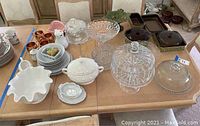 Wide view of table displaying multiple ceramic and glass items including cake stands, plates, and small bowls along with copper mugs in background.