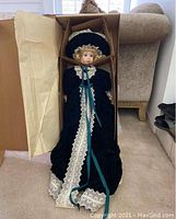 Porcelain doll packaged in a box with cardboard supports, dressed in a dark green velvet Victorian-style dress with lace and ribbons.