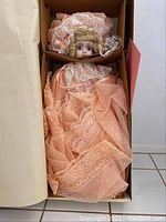 Porcelain doll lying in original box, showing upper body, bonnet, and details of peach lace dress.