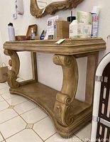 Decorative wood entry table shown from left angle, showcasing the scrollwork legs, carved details along the sides and bottom shelf, and various items placed on the top surface.
