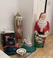 Santa Claus figure, tree stands, holiday plate, light decoration, and other small holiday decor items on the floor near a wall