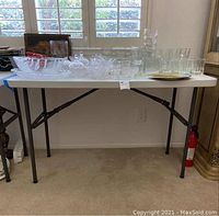 White Staples branded plastic folding table with black metal legs. Shows some scratches on tabletop surface. Various glassware items placed on top, not included in the lot.