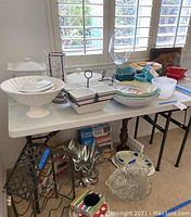 General photo showing white folding plastic table with household items on and under the table near the window with wooden blinds.