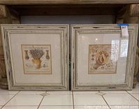 Two framed shabby chic style prints resting against a piece of furniture. Both frames are white distressed wood measuring about 23x23 inches each.