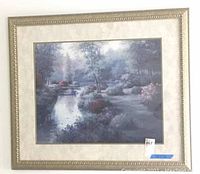 Front view of framed landscape painting hung on a wall showing serene river scene with trees and flowers
