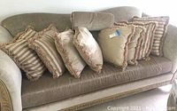 Lot of 9 decorative pillows displayed on a couch, showing various designs and colors mostly in beige and taupe with fringe borders.
