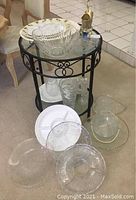 Various clear glass plates, vases, and trays arranged on a small black metal and glass round side table and surrounding area. Items include a large decorative bowl on top, two vases inside the table's shelves, and multiple plates and trays on the floor.