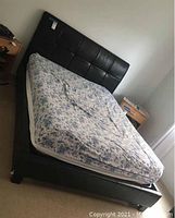 Full view of bed with tufted black faux leather backboard and mattress covered with blue floral sheet.