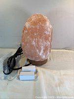 Large pink-orange Himalayan salt lamp approximately 9 inches tall on wooden base with attached electrical cord and two small white boxes.