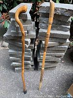 Full view of two wooden canes set against stacked bricks. One cane has a curved handle and the other is a carved walking stick.