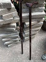 Full view of two 36-inch tall wooden canes with carved straight handles and rubber footings placed against a stone background.