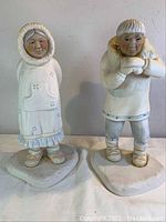 Front view of two porcelain statues depicting Alaskan indigenous figures, one female and one male, standing on flat sculpted bases.