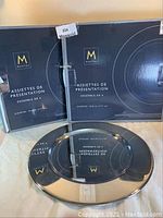 Two black original boxes labeled Mantle with 4 charger plates each, and 2 silver plated charger plates displayed in front of boxes.