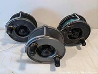 Front view of three vintage fishing reels showing metal bodies, black handles and grips, with fishing line on two reels.