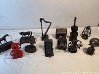 Photo showing all 12 miniature metal pencil sharpeners arranged in a row. Red fire hydrant and multiple bronze-colored items including horses, harp, violin, lantern, globe, fans, and machines.